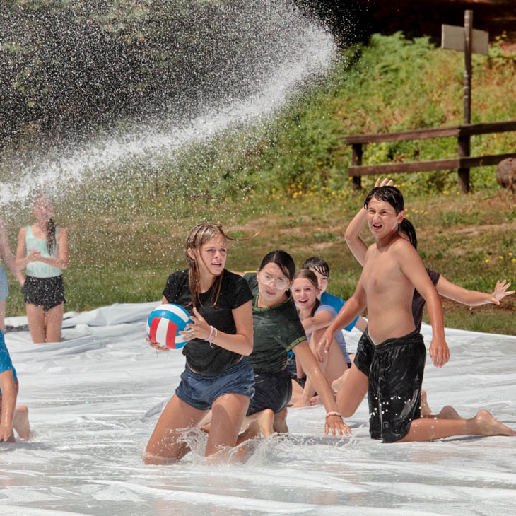 Children playing in a shallow water area, chasing a volleyball as water splashes around them under a spray of water.