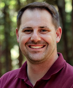 Smiling man in a burgundy polo shirt outdoors in a forest setting.