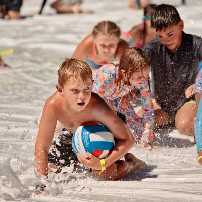 2026-7-28 Kids Summer Camp-19 Children play in soapy foam, crawling on the ground. One boy in front clutches a beach ball, looking determined. Other kids, wet and smiling, follow behind him, all wearing swimsuits and wristbands.