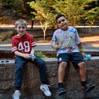 2026-7-28 Kids Summer Camp-38 Two boys sit on a low stone wall outdoors, holding blue ice cream. One wears a red 48 shirt and dark pants; the other wears a colorful shirt and shorts. Green trees are in the background.