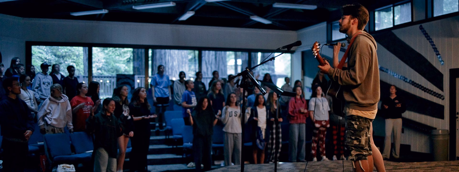 2027 Program Banner A man plays guitar and sings into a microphone on stage while a group of people stands and listens in a room with large windows and trees visible outside.