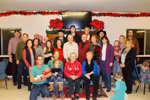 Close up staff photo A group of people of various ages pose together indoors, dressed in casual winter clothing, with holiday decorations and poinsettias in the background. Two elderly women are seated in front, surrounded by family members.