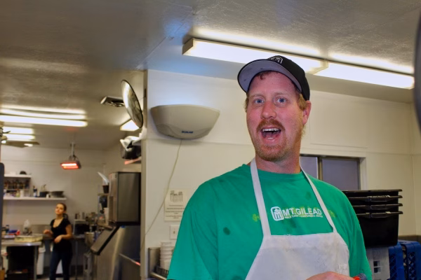 Alumni Pictures 11 A man in a green shirt and white apron smiles in a kitchen, wearing a black cap. Another person stands in the background near kitchen equipment.