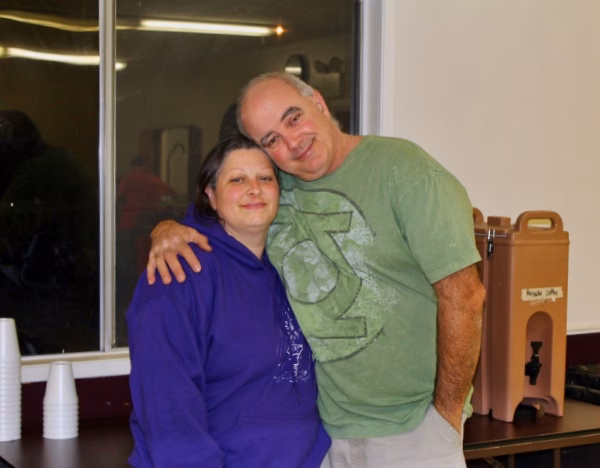 Alumni Pictures 12 Two people stand indoors, smiling and hugging. The person on the left wears a purple hoodie; the person on the right wears a green shirt. Disposable cups and a beverage dispenser are visible nearby.