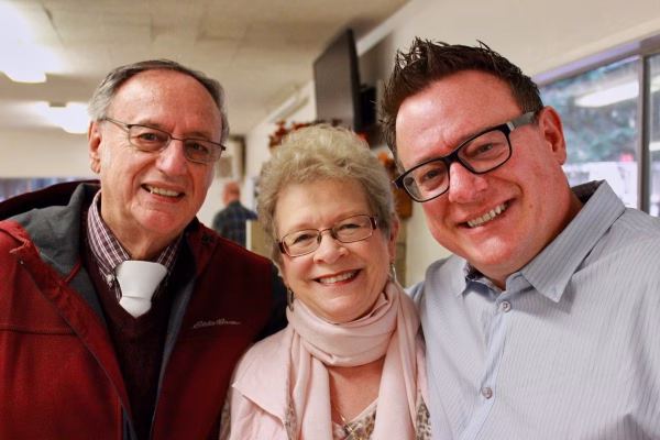 Alumni Pictures 13 Three smiling adults pose together indoors. The man on the left wears glasses and a red jacket, the woman in the center has short hair and a pink scarf, and the man on the right wears glasses and a light blue shirt.