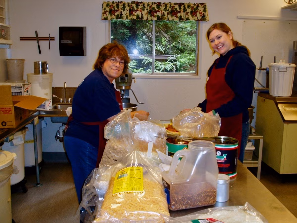 OLYMPUS DIGITAL CAMERA Two women in aprons prepare food at a kitchen counter, surrounded by bulk food items like oats and spices. They are smiling and working together in a well-lit kitchen with a window in the background.