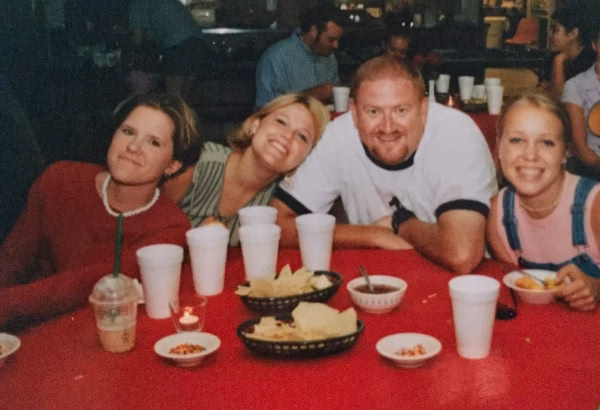 Four people sit at a red table with baskets of chips, bowls of salsa, and cups. They are smiling and leaning together, appearing happy and relaxed in a casual dining setting.