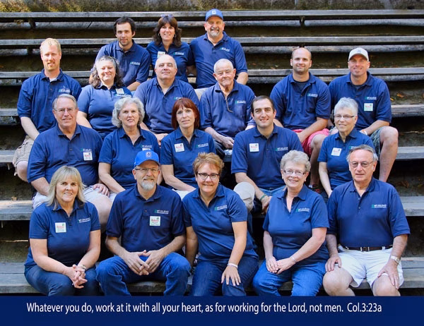 PRETTY GOOD SHOT YEAR ROUND STAFF A group of nineteen adults in matching blue shirts sit on outdoor steps, smiling at the camera. A Bible verse is displayed at the bottom of the image.