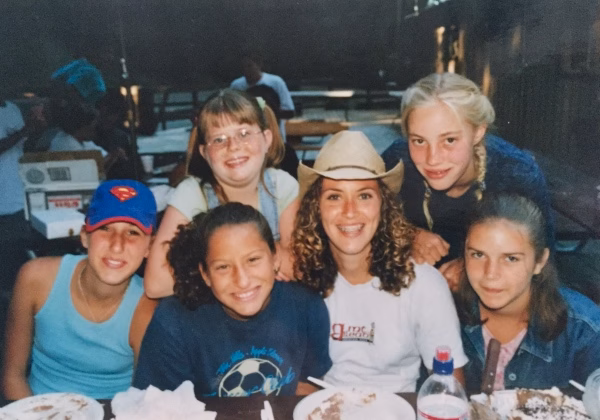 Alumni Pictures 22 Six smiling girls, some with braided hair and glasses, sit closely together at a table with plates of food. One girl wears a cowboy hat, and another wears a blue tank top and a red Superman cap. They appear to be at an outdoor gathering.