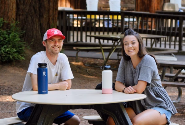 Alumni Pictures 4 Two people sit outside at a round picnic table, smiling at the camera. Each has a water bottle on the table. Benches and trees are visible in the background.
