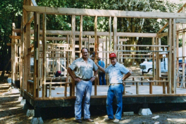 Alumni Pictures 5 Two men stand and smile in front of a wooden house frame under construction in a wooded outdoor setting, with other people and a vehicle visible in the background.