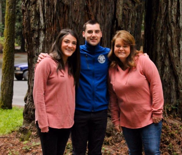 Alumni Pictures 6 Three people smile and pose in front of a large tree. Two women wear pink hoodies, and a man in a blue jacket stands between them, with his arms around both. Lush greenery and a road are visible in the background.