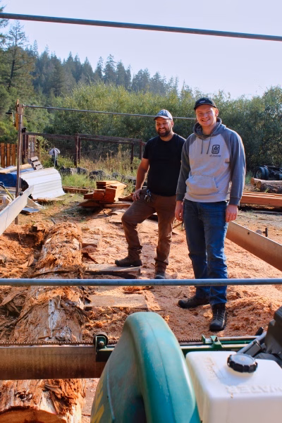 Alumni Pictures 8 Two men stand by a large log and sawmill outdoors, surrounded by wood and trees. Both are smiling and dressed in casual work clothes, with forested hills in the background.