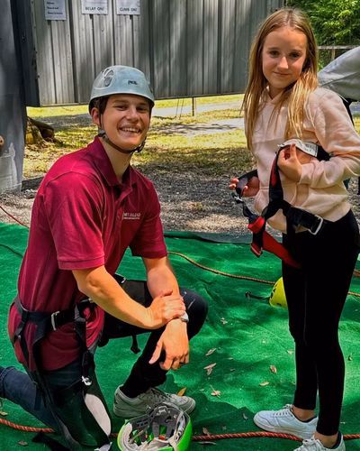 A man in a helmet and maroon shirt kneels on green turf next to a smiling girl holding a climbing harness, with trees and a building in the background.