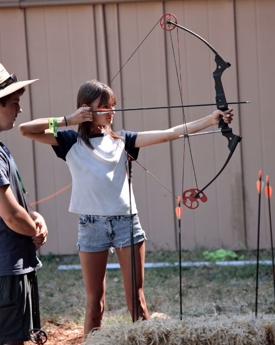 A girl in a white and black shirt aims a compound bow at an outdoor archery range while a person in a hat stands nearby. Arrows and hay bales are visible in the background.