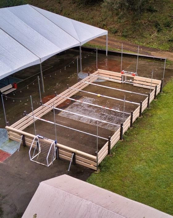 An outdoor fenced sports court with two soccer goals, surrounded by grass and covered partially by a large white tent. The surface appears wet, likely after rain.