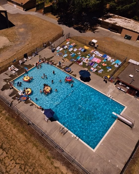 Aerial view of a rectangular outdoor swimming pool with people swimming and lounging on floats; towels and umbrellas are laid out around the pool on concrete.