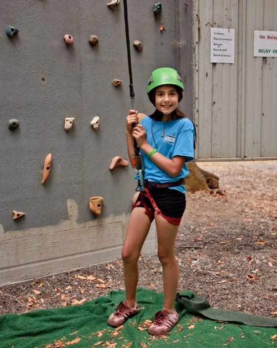 A smiling girl wearing a helmet and harness stands by a climbing wall, holding a safety rope. She is on a green mat, ready to climb, with fallen leaves and climbing grips visible around her.