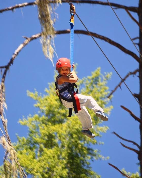 A child wearing a red helmet and harness smiles while zip-lining outdoors against a backdrop of green trees and a clear blue sky.