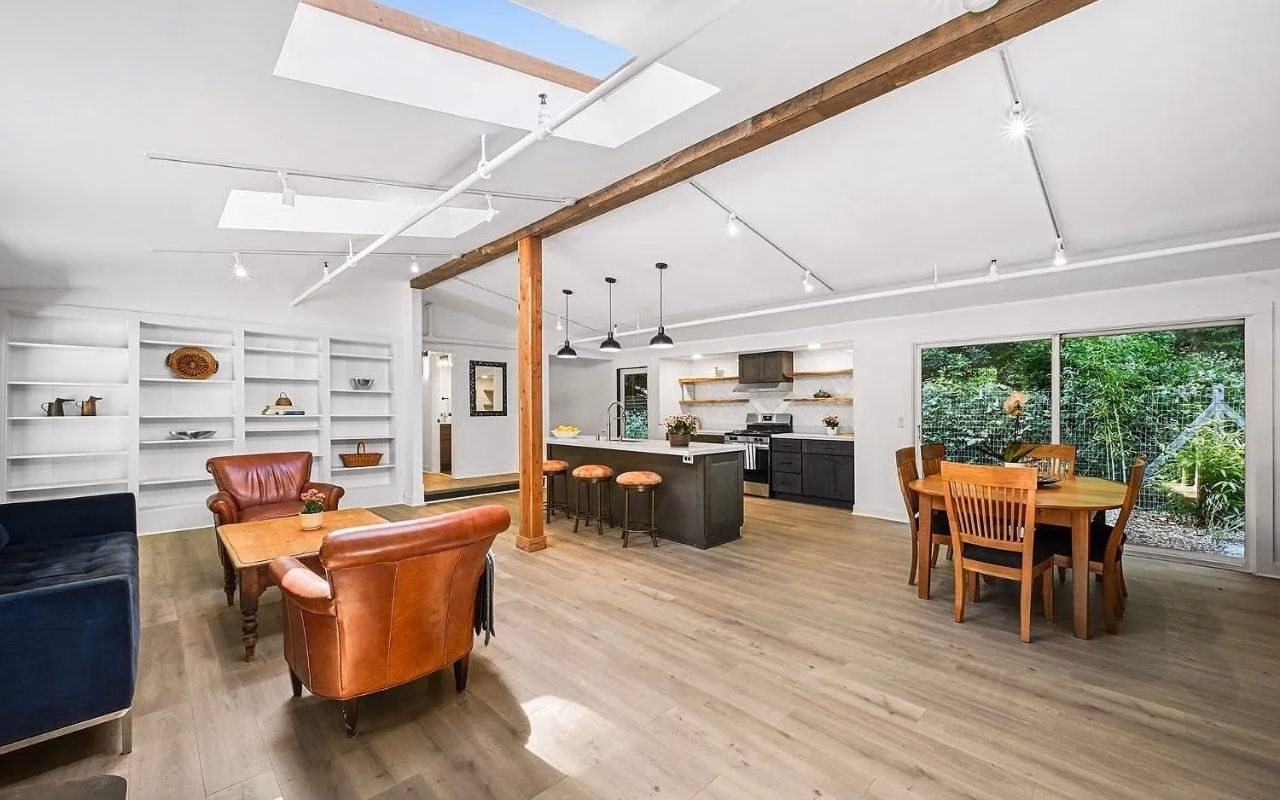 Modern open-concept living space with leather chairs, built-in shelves, a kitchen with barstools, dining table, wood floors, skylights, and large windows overlooking a green backyard.