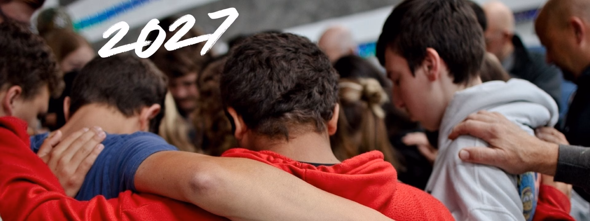 Youth Winter Retreat Banner 2027 v3 A group of people stand close together with arms around each other, heads bowed. The number 2027 is written in white text in the top left corner.