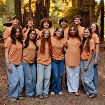 tim-teams-feature-1 2027 Twelve smiling teens in orange shirts and blue jeans stand close together in a forest, posing for a group photo with tall trees in the background.
