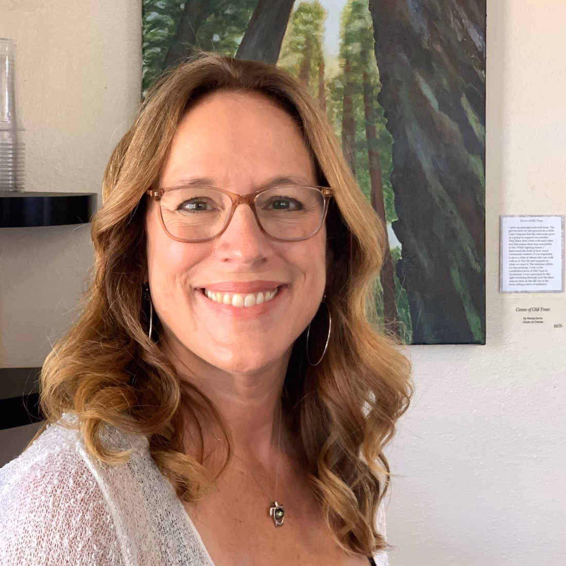 IMG_3330 SQUARE A smiling woman with wavy light brown hair and glasses stands in front of a painting of trees.