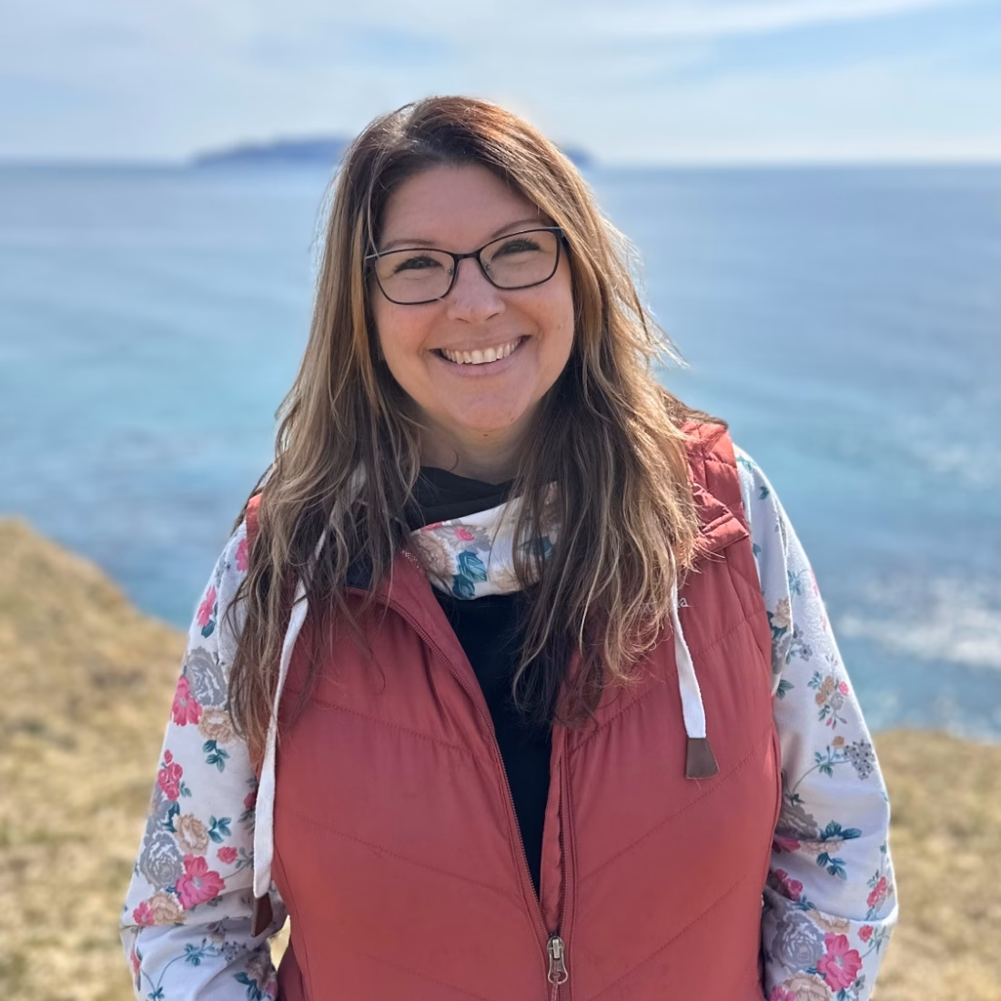 Terah Lites SQUARE A woman with long hair and glasses smiles while standing in front of an ocean.